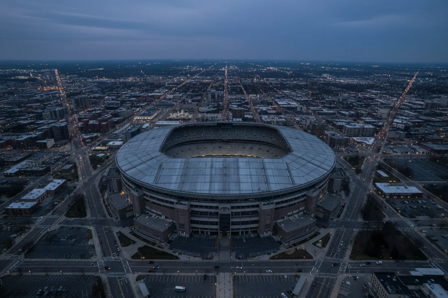 An oblique aerial view of a stadium exterior and the surrounding city grid at dusk, with the horizon and sky across the top of the frame