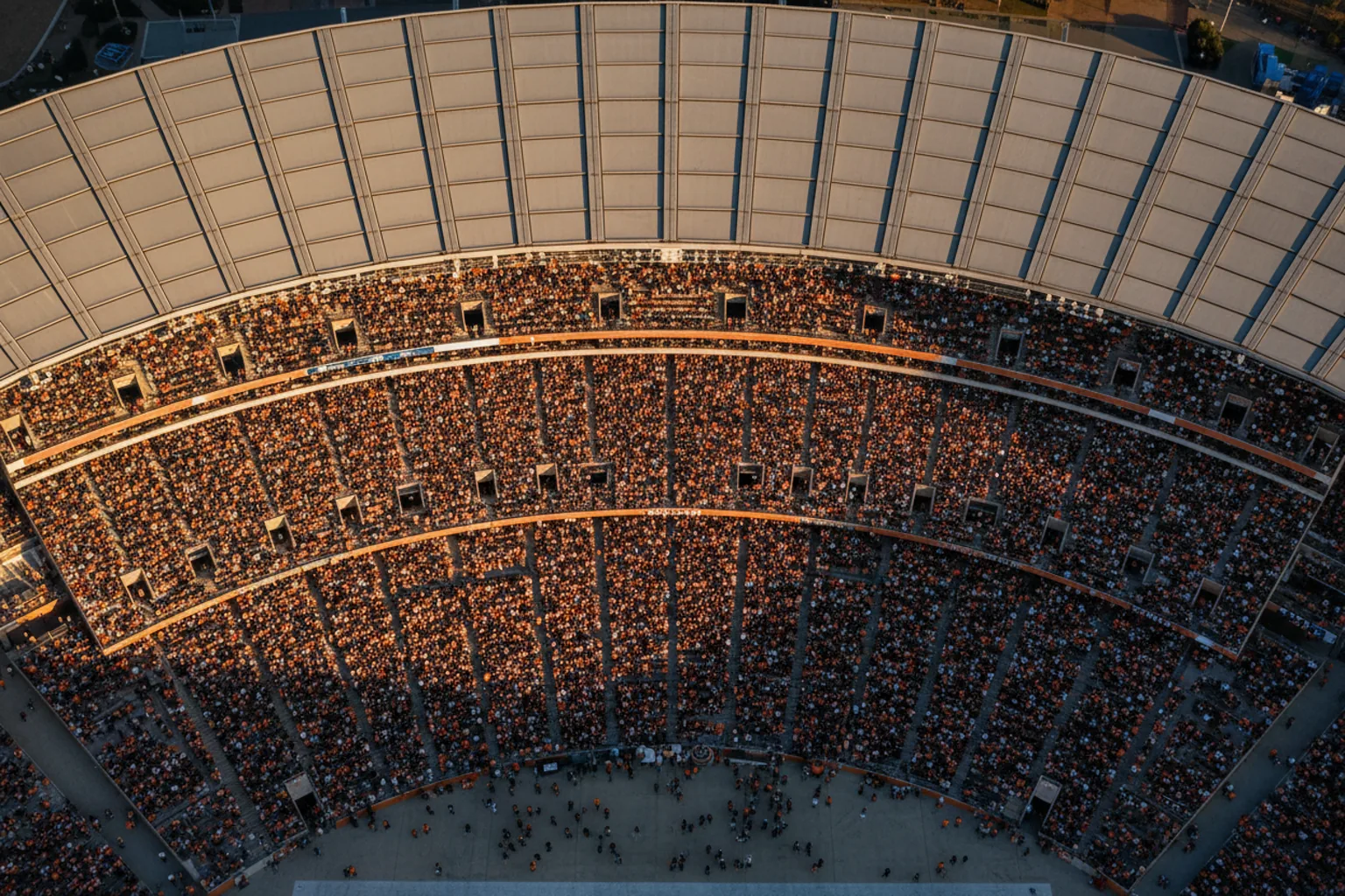 Aerial view along the curved rim of a packed stadium at golden hour, a brown-and-orange crowd filling the tiers