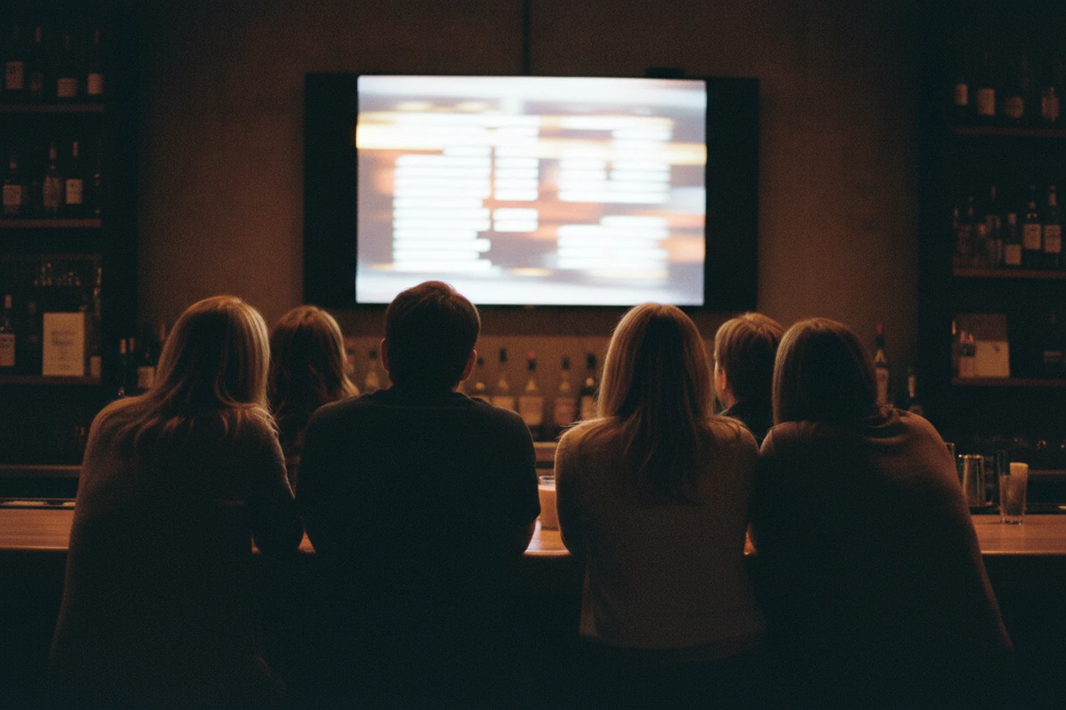 Six people seen from behind at a dimly lit bar, all watching a wall-mounted screen that shows an indistinct grid of pale rows