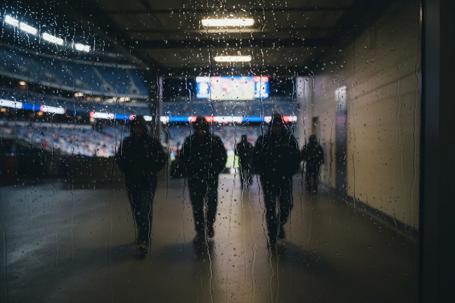 A rain-streaked window at night with the navy-and-red glow of a stadium beyond the glass